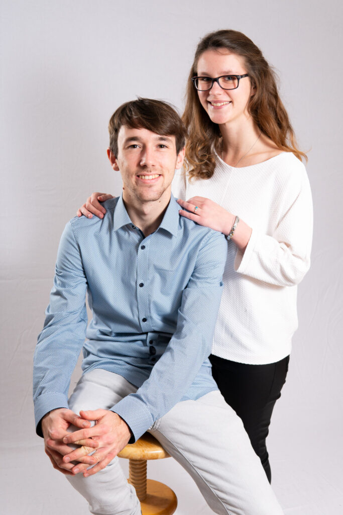 Junger Mann in blauem Hemd sitzt auf Stuhl neben junger stehender Frau, die sich an ihn lehnt